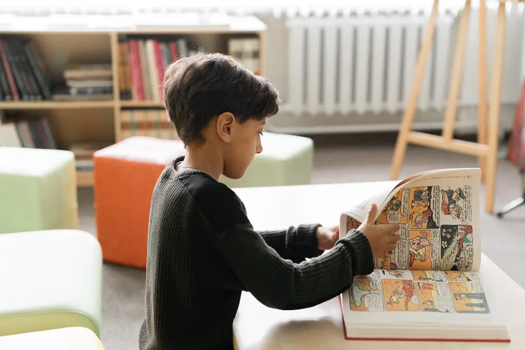 A boy reading colorful comic books at a table in a cozy library room Image depicting books.
