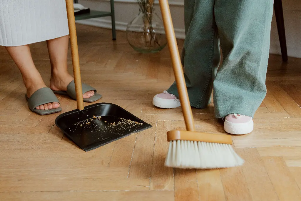 Two people using a broom and dustpan to clean floor. Image depicting dustpan.